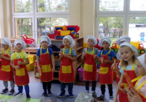 Dzieci stojące na dywanie w strojach kucharzy, trzymają marchewki przygotowane do mycia.